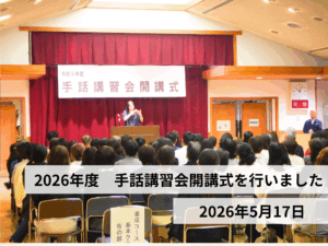 会場の前方ステージに『令和8年度 手話講習会 開講式』の横断幕。客席に参加者が座り、演台前で手話表現をしている人がいる。