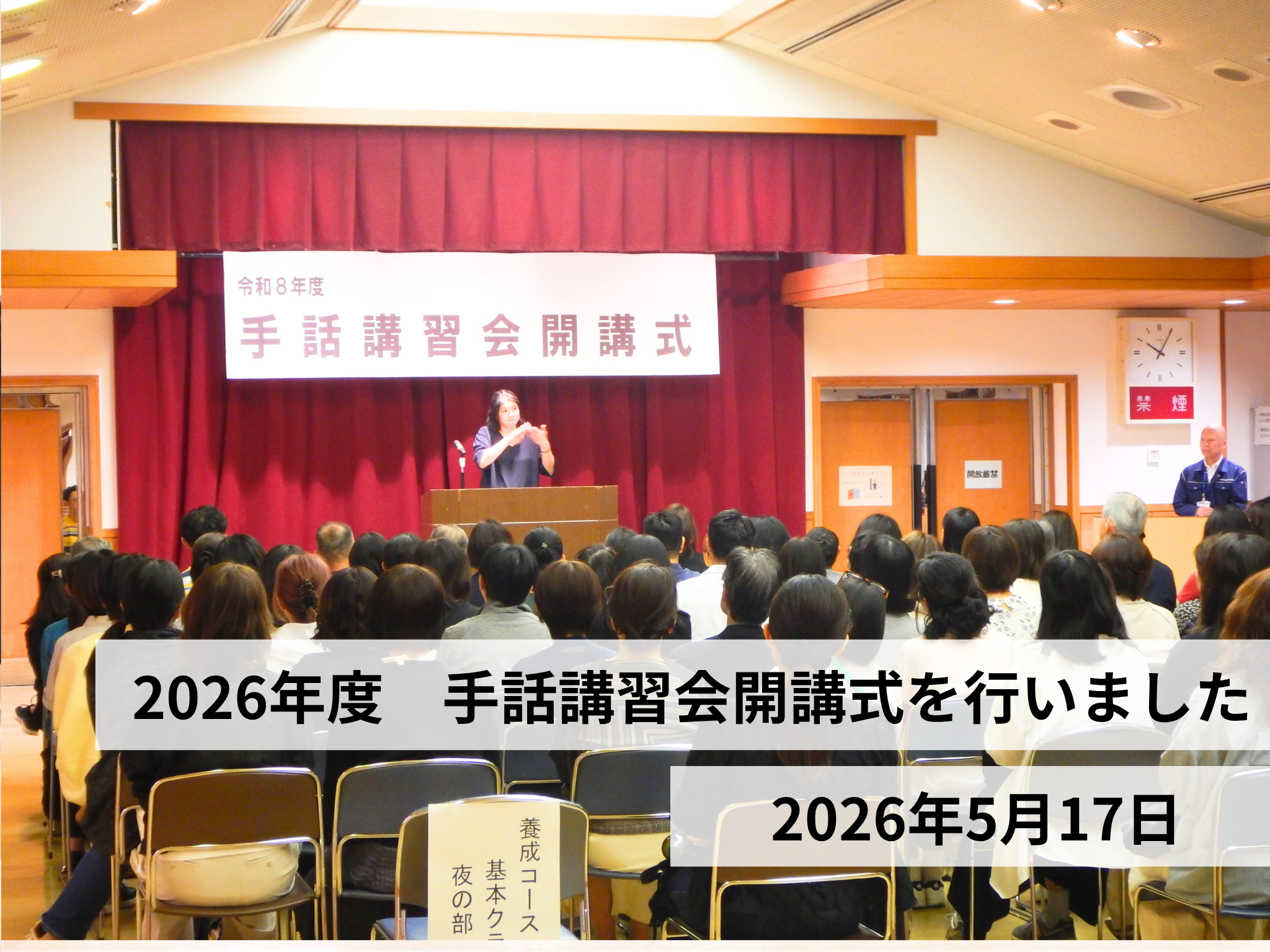 会場の前方ステージに『令和8年度 手話講習会 開講式』の横断幕。客席に参加者が座り、演台前で手話表現をしている人がいる。