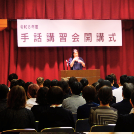 会場の前方ステージに『令和8年度 手話講習会 開講式』の横断幕。客席に参加者が座り、演台前で手話表現をしている人がいる。